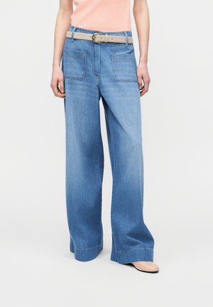 Person trägt hellblaue Jeans mit weitem Bein und großen Fronttaschen, beigen Gürtel, pfirsichfarbenes Oberteil und beige Schuhe.