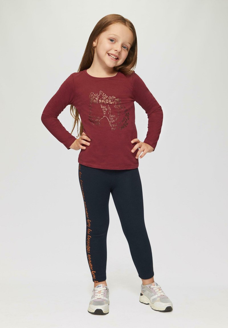 Bordeaux langemouwen shirt met grafisch ontwerp, gecombineerd met marineblauwe legging met oranje tekst aan de zijkant. Getextureerde sneakers maken de outfit compleet.