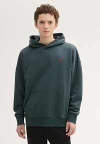 Ciemnozielona bluza z kapturem z bawełny, z przednią kieszenią, w luźnym fasonie, z małym czerwonym logo na piersi. Miękka tekstura, casualowy design.