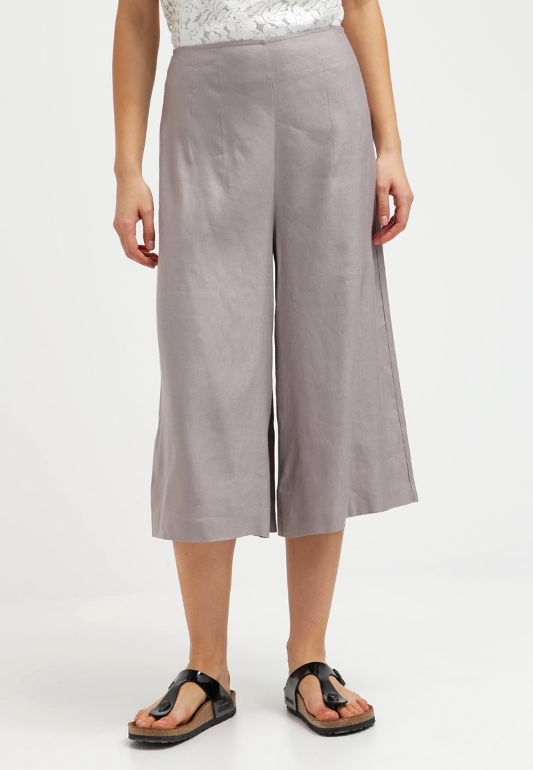 Femme portant un pantalon gris taille haute, jambes larges et court, associé à des sandales noires et un haut en dentelle blanche, debout sur un fond blanc uni.