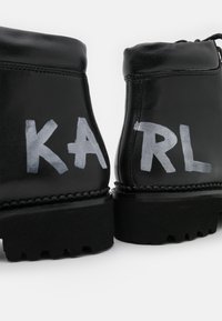 Stivaletti neri in pelle con suole in gomma spesse, con lettere bianche "KARL" dipinte a mano sul retro, finitura testurizzata, design minimalista.