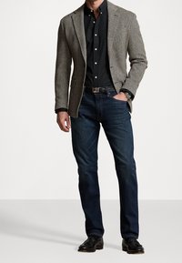 Gråbrun kavaj med fiskbensmönster, bärs över en svart knappskjorta. Mörkblå jeans och svarta skor fulländar outfiten.