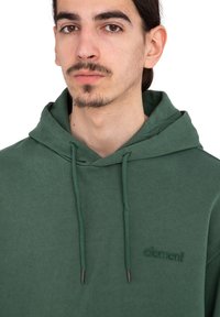Joven con cabello oscuro y vello facial, vestido con una sudadera con capucha verde oscura con cordones y un pequeño logo de "element" en el pecho.