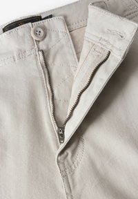 Los pantalones de color beige claro cuentan con un cierre de cremallera metálica y un botón en la parte superior. La tela es suave y tiene sutiles detalles de costura cerca de la cremallera.