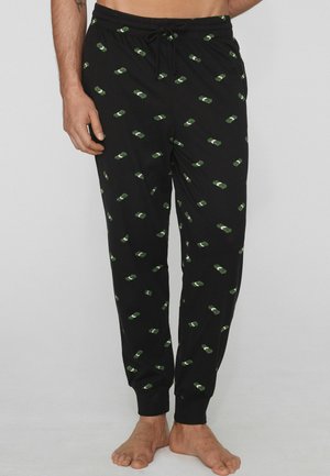 Hombre vistiendo pantalones deportivos negros con pequeños estampados verdes de fajos de dinero, descalzo, de pie frente a un fondo liso y claro.