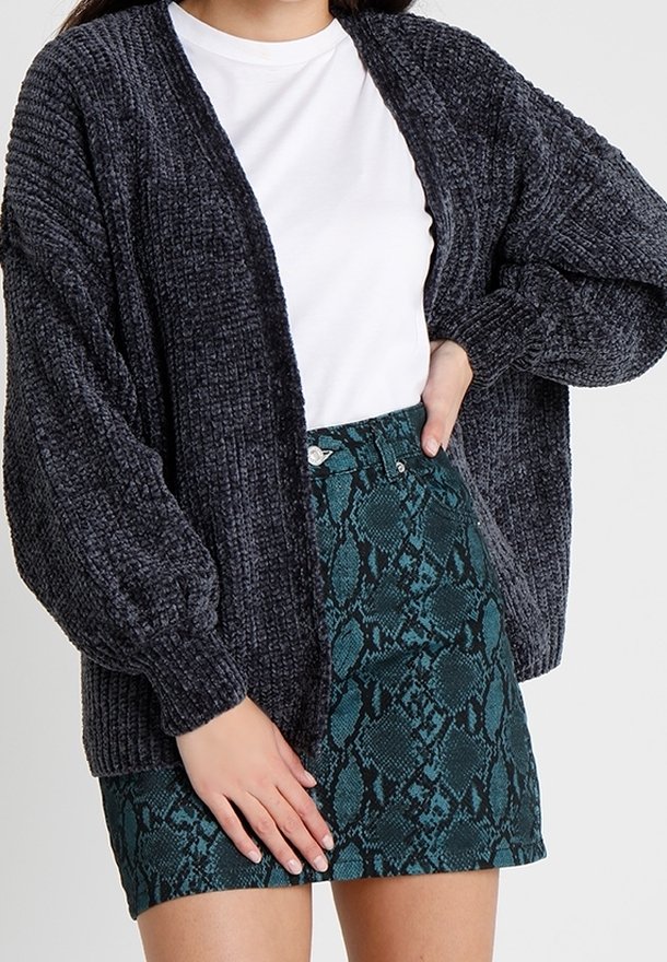 Femme portant un cardigan en maille épaisse gris foncé, un t-shirt blanc et une mini-jupe verte à motif peau de serpent, debout avec une main sur la hanche.