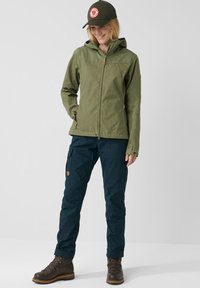 Olivgrön zip-up jacka med huva, tillsammans med mörkblå byxor med cargofickor. Bruna snörstövlar och en grön keps.