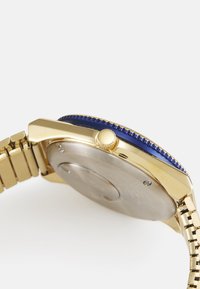 Montre-bracelet dorée avec une lunette rotative bleue, une couronne texturée et un bracelet en acier inoxydable. Elle présente un dos lisse et métallique.