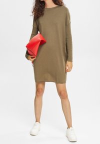 Abito in maglia verde oliva con maniche lunghe e vestibilità ampia, abbinato a una borsa a tracolla rossa e sneakers bianche. Texture liscia, design minimale.