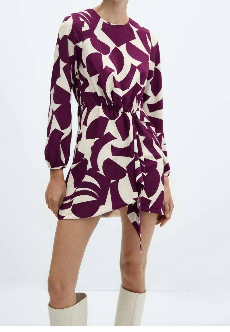 Femme portant une robe mini à manches longues, à motifs abstraits violets et blancs, avec une ceinture à la taille, assortie à des bottes blanches jusqu'aux genoux.