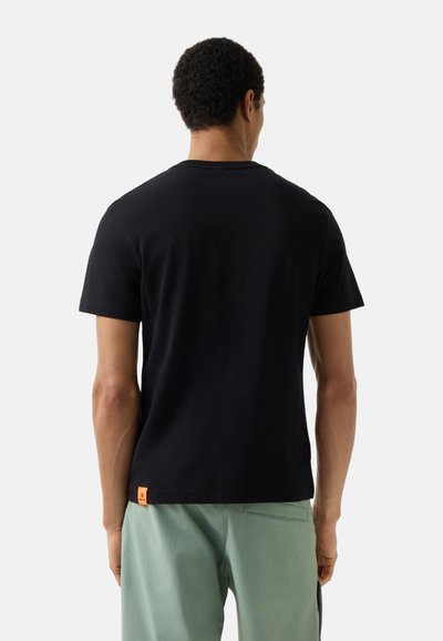 Muž nosící jednoduché černé tričko s krátkým rukávem a světle zelené kalhoty, stojící zády k divákovi před jednoduchým pozadím.