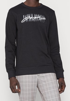 Sudadera negra de manga larga, con cuello y puños acanalados; presenta un diseño gráfico en texto blanco en la parte delantera. Se lleva con pantalones de cuadros.