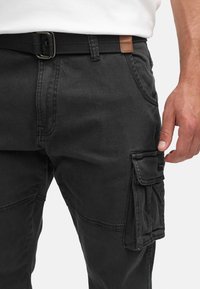 Homme portant un pantalon cargo noir avec une poche latérale, une ceinture noire et une chemise blanche, la main détendue près de la poche.