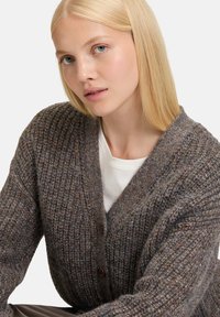 Harmaa neulecardigan, jossa on tekstuuri, syvällä V-pääntiellä, suurilla nappeilla ja rentolla istuvuudella. Päällä valkoinen paita.