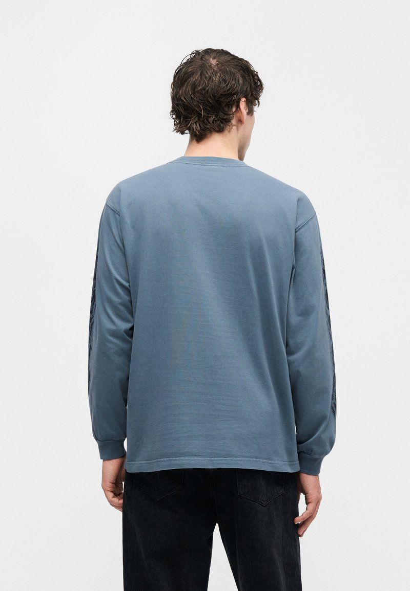 Mann mit lockigem braunem Haar, der ein blaues langärmeliges Hemd und schwarze Hose trägt, steht mit dem Rücken zur Kamera vor einem schlichten Hintergrund.