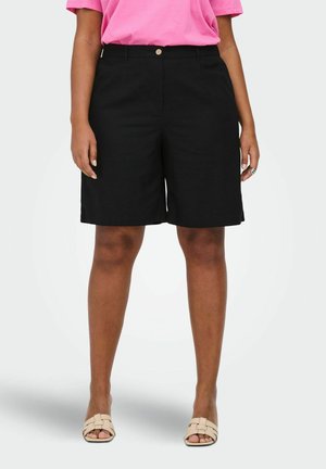 Femme portant un short noir mi-long, un haut rose à manches courtes, des sandales beiges tressées ouvertes aux orteils, et des bagues argentées à la main droite.