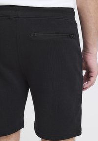 Pantalones cortos negros con una textura suave, cintura elástica y un bolsillo con cremallera en la parte trasera. El dobladillo es recto y termina por encima de la rodilla.