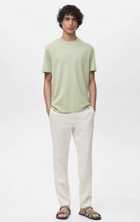 Světle zelené tričko z jemné látky, spárované s krémovými lněnými kalhotami. Model nosí hnědé sandály na pásky a stojí na jednobarevném pozadí.