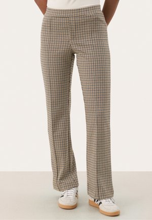 Unterkörper mit beige-schwarz karierten, geraden Hosen, kombiniert mit weißen Turnschuhen mit schwarzen Streifen und braunen Sohlen.