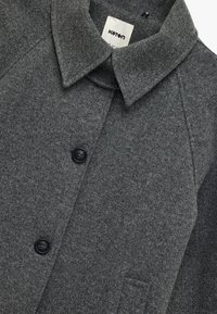 Abrigo de lana gris con un cuello clásico, que presenta botones negros y un bolsillo de parche, destacando un tejido texturizado y un diseño a medida.