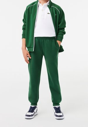 Survêtement de sport vert avec des accents blancs, comprenant une veste zippée, un pantalon à taille élastique et des baskets bleu marine. Polo en dessous.