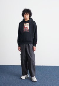 Sudadera negra con un gráfico frontal, combinada con pantalones cargo grises. El modelo lleva zapatillas blancas y está de pie sobre una alfombra azul en un fondo sencillo.
