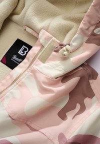 Jachetă de camuflaj roz ușoară, cu căptușeală moale din fleece bej, dotată cu fermoar și șnururi ajustabile. Eticheta brandului este vizibilă în interior.