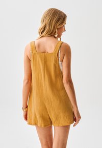 Senffarbener Jumpsuit aus strukturiertem Stoff mit breiten Trägern, weiter Passform und kurzen Hosenbeinen. Keine sichtbaren Verschlüsse oder Muster.