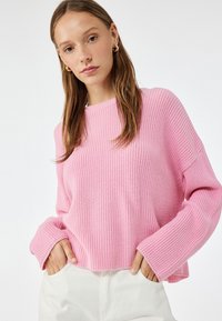 Maglione rosa a costine con ampi polsini e scollo rotondo. Indossato con pantaloni bianchi, che mettono in mostra una vestibilità rilassata e un tessuto testurizzato.