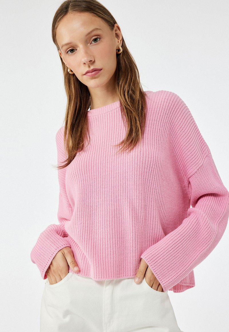 Maglione rosa a costine con ampi polsini e scollo rotondo. Indossato con pantaloni bianchi, che mettono in mostra una vestibilità rilassata e un tessuto testurizzato.