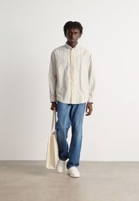 Licht gestreept overhemd in beige met blauwe en groene strepen, blauwe denimbroek en witte sneakers; draagt een crèmekleurige tote bag.