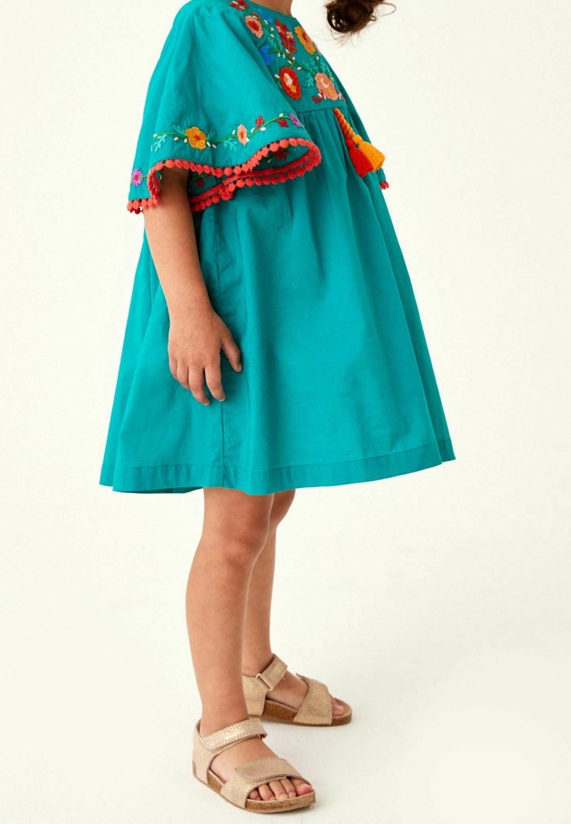 Robe en coton turquoise avec des motifs floraux brodés, des franges en pompons et une silhouette évasée, assortie à des sandales beiges.