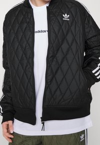 Svart vadderad bomberjacka med dragkedja, ribbade muddar och nederkant. Vit Adidas-logotyp på bröstet och axeln, tillsammans med en vit t-shirt under.