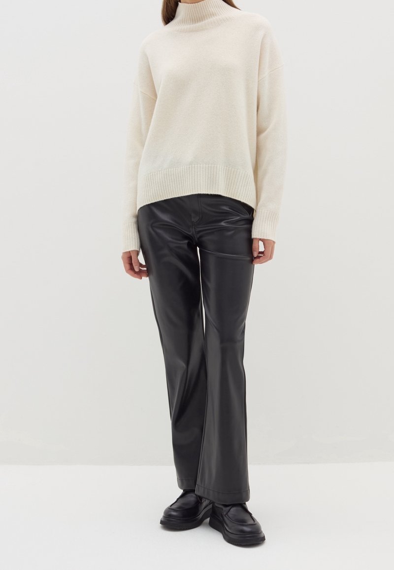 Femme portant un pull à col roulé crème, un pantalon droit en cuir noir et des mocassins noirs épais, debout sur un fond blanc.