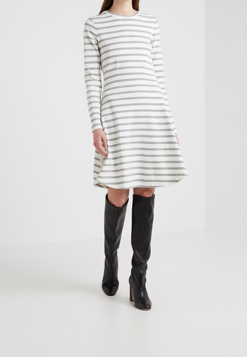 Femme portant une robe à manches longues blanche et grise à rayures horizontales, associée à des bottes en cuir noires hautes jusqu'aux genoux, debout sur un sol blanc.