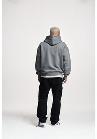 Sudadera gris de gran tamaño con capucha ajustable, puños y dobladillo acanalados, combinada con pantalones cargo negros que tienen bolsillos laterales. Fondo liso.
