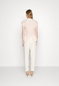 Blusa rosa chiaro con maniche corte e orlo curvo, abbinata a pantaloni bianchi e tacchi beige, vista da dietro.