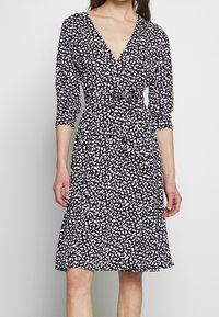 Czarna sukienka z białym wzorem liści, dekolt w kształcie litery V, rękawy o długości trzech ćwierci, dopasowana w talii i zwiewny dół. Lekki materiał o miękkiej teksturze.