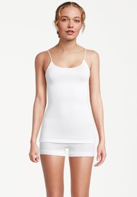 Donna che indossa un top senza maniche bianco aderente e uno slip boyshort bianco abbinato, in piedi contro uno sfondo bianco semplice.