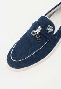 Navyblauwe denim loafers met een witte leren bies, een zilveren sleutelhanger en een geborduurd logo op de bovenkant. Glad hakje met subtiele textuur.