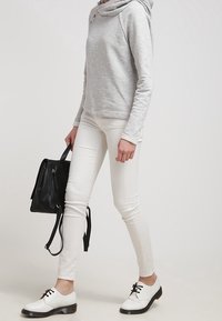 Persona con un top grigio a maniche lunghe, pantaloni bianchi e scarpe bianche, che tiene una borsa in pelle nera su uno sfondo neutro.