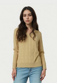 Pulover tricotat cu model în relief, de culoare galben deschis, cu mâneci lungi și manșete canaletate, având un mic emblemă albă pe piept. Asortat cu blugi albaștri.