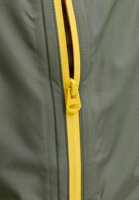 Detaljni prikaz žutog patentnog zatvarača djelomično otkopčanog na maslinasto zelenom tkanju, prikazujući detaljno zube patentnog zatvarača i povlačnu traku.