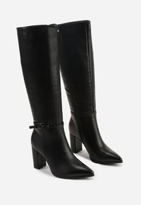 Bottes noires en cuir montantes jusqu'au genou avec un bout pointu et un talon carré. Elles sont dotées d'une fermeture éclair sur le côté et d'une boucle décorative autour de la cheville.