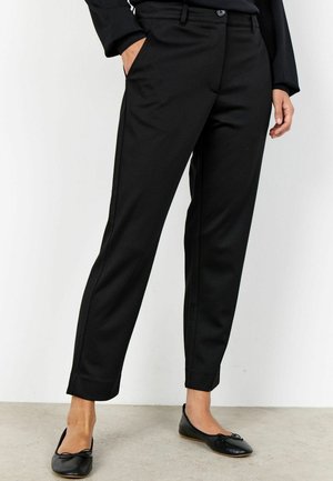 Kobieta ubrana w dopasowane czarne spodnie do kostek oraz czarne baletki, stojąca z jedną ręką w kieszeni na białym tle.