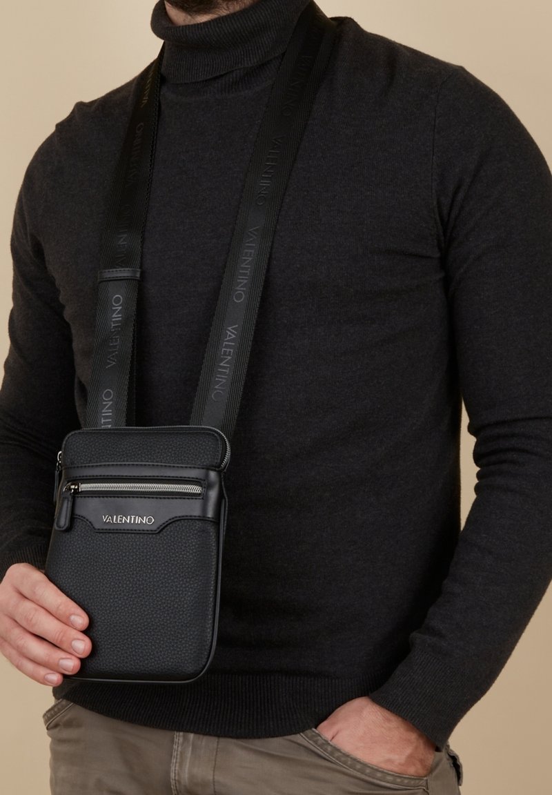 Borsa a tracolla nera con finitura testurizzata, tasca con zip e tracolla regolabile con logo del marchio ripetuto. Tenuta da una persona in un maglione scuro.