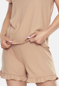 Osoba poprawia dół jasnobrązowego, prążkowanego topu i dopasowanych szortów, prezentując swobodny styl z falbaniastymi brzegami.