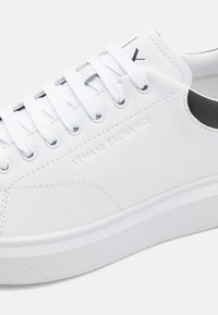 Sneaker bianca in pelle con lacci piatti, caratterizzata da un accento nero sul tallone e il logo "Armani Exchange" in rilievo sul lato.