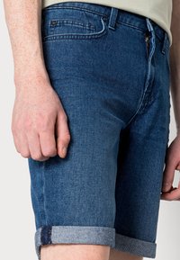 Shorts en denim bleu foncé avec un ourlet roulé, dotés d'une fermeture éclair, de poches et d'un tissu lisse et texturé. Main reposant sur la cuisse.