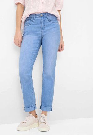 Frau trägt hellblaue umgeschlagene Jeans, blassrosa strukturierte Bluse und beige Schnürsneaker, steht vor weißem Hintergrund.